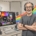 Smiling middle-aged male video creator in gray tee-shirt with headphones on. Computer screen with colorful images. In home studio with video lights. Splash of rainbow colors across background.